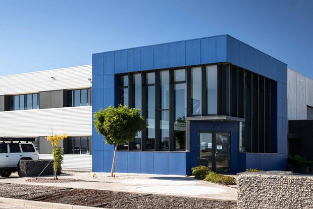 Exterior view of a contemporary low-rise commercial building with a bold blue metal-clad entrance volume, tall dark-framed glazed facade, and adjacent white horizontal cladding. The building fronts a paved entry with sparse landscaped beds and textured stone retaining walls under clear daylight.