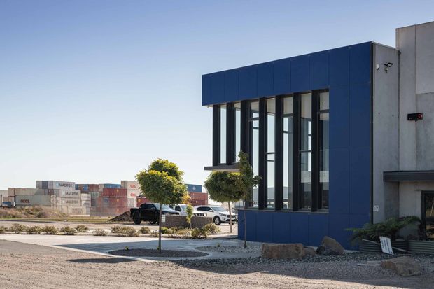 Exterior view of a contemporary low-rise commercial or industrial building with a dark blue metal-panel facade, tall black-framed glazed openings, and exposed concrete side walls. The building sits within a paved and gravelled landscaped site with small trimmed trees and rocks. Shipping containers and parked vehicles are visible in the industrial surroundings.
