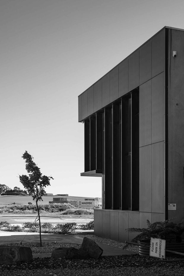 Black-and-white exterior photograph of a contemporary commercial or industrial building. The facade is composed of dark metal or fibre-cement-like panels with deep vertical recessed openings and projecting horizontal planes. A landscaped foreground with rocks, gravel and a young tree leads toward the building, while an industrial shipping-container setting is visible in the background.