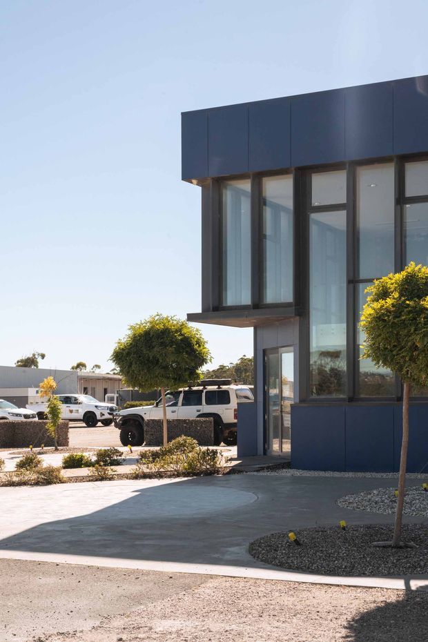 Exterior view of a contemporary two-storey building with a dark blue, box-like facade, extensive dark-framed glazing, and projecting upper-level volumes. The building fronts a paved landscaped forecourt with small clipped trees, gravel garden beds, and low shrubs. Vehicles and a low industrial-looking structure are visible in the background.