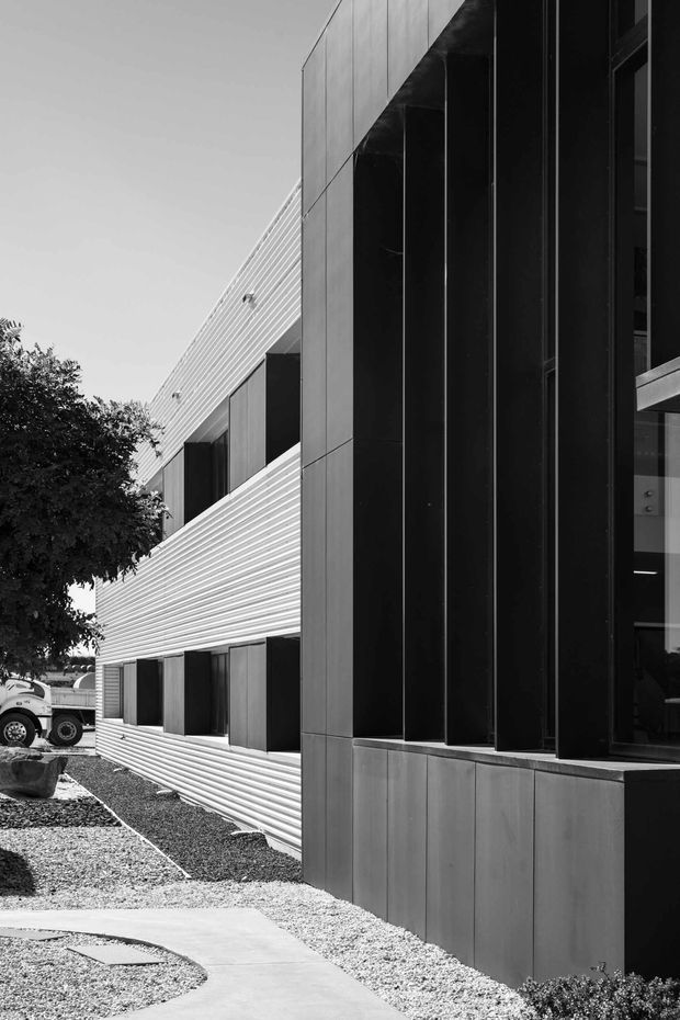 Black-and-white architectural photograph of a contemporary multi-storey building exterior. The facade combines horizontal ribbed cladding with dark vertical fins and large recessed windows, alongside a gravel landscape and curved concrete walkway.
