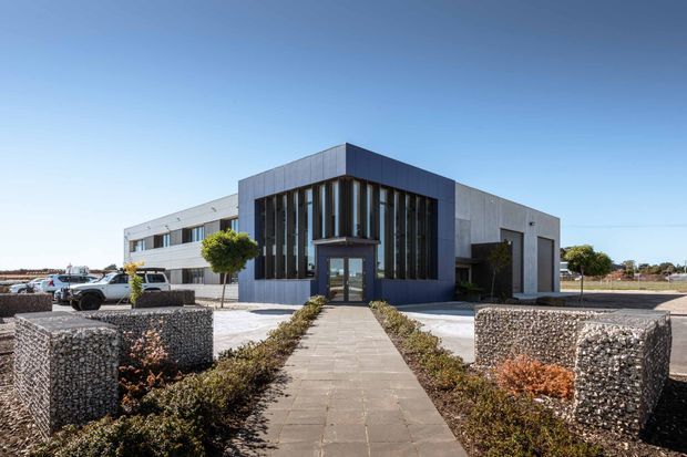 Contemporary low-rise commercial building viewed from the front, with a dark charcoal-clad entrance volume, tall narrow vertical glazing, and pale rendered or panelled wings. A paved walkway leads through minimalist landscaping with gabion retaining walls, clipped trees, and parking visible to the side.
