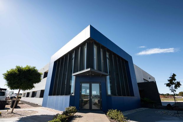 Exterior view of a contemporary commercial building with a sharply faceted, angular form. The dark blue and grey facade combines extensive vertical glazing with solid metal-clad volumes, creating a prominent glazed entrance beneath a projecting canopy. Low landscaping and paved paths frame the building under a clear sky.