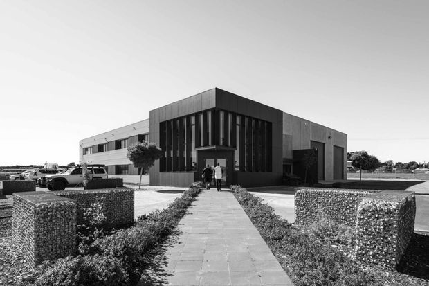 Black-and-white exterior photograph of a contemporary low-rise commercial or institutional building. The composition centres on a paved pedestrian path leading through clipped hedge and gabion-style landscape walls to a recessed entrance. The building has a strongly articulated dark central volume with tall vertical glazing and fins, flanked by lighter, horizontally composed wings with flat roofs. Parking and additional low-rise structures are visible in the background.