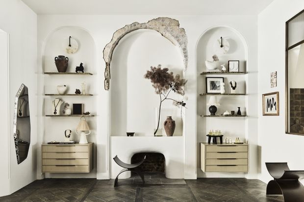 Refined living room or entry display area featuring three tall arched wall niches, a central exposed masonry arch over a recessed fireplace, floating shelves with curated objects, and symmetrical built-in drawer units. The space has white plaster walls, dark herringbone timber flooring, sculptural black furniture, and a large black-framed window or glazed opening. The overall look is curated, historic-inspired, and contemporary.