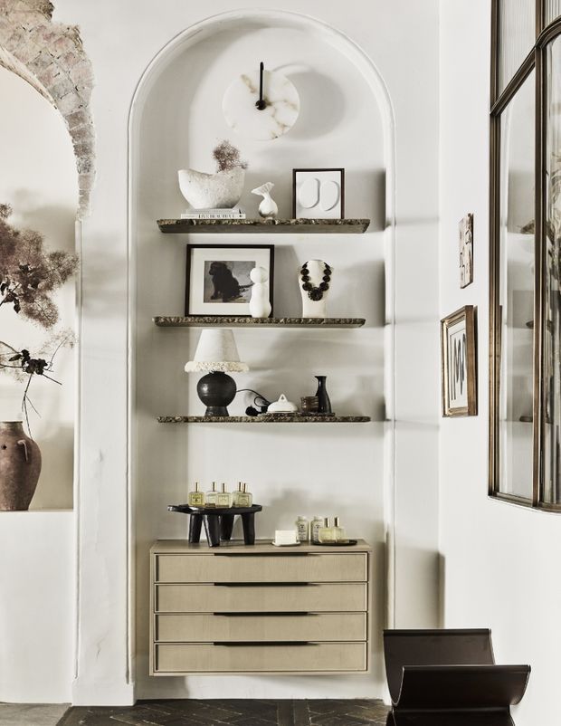 A refined interior display niche with a tall arched plaster recess, three dark stone or marble shelves, curated ceramics, framed artwork, decorative objects and a clock. A low beige built-in or fitted chest of drawers sits beneath the shelving, with a small side table and chair nearby. The surrounding walls are white with an exposed aged masonry arch at left, while a large dark-framed mirror reflects the display. The room has a warm, monochromatic, artfully styled character.