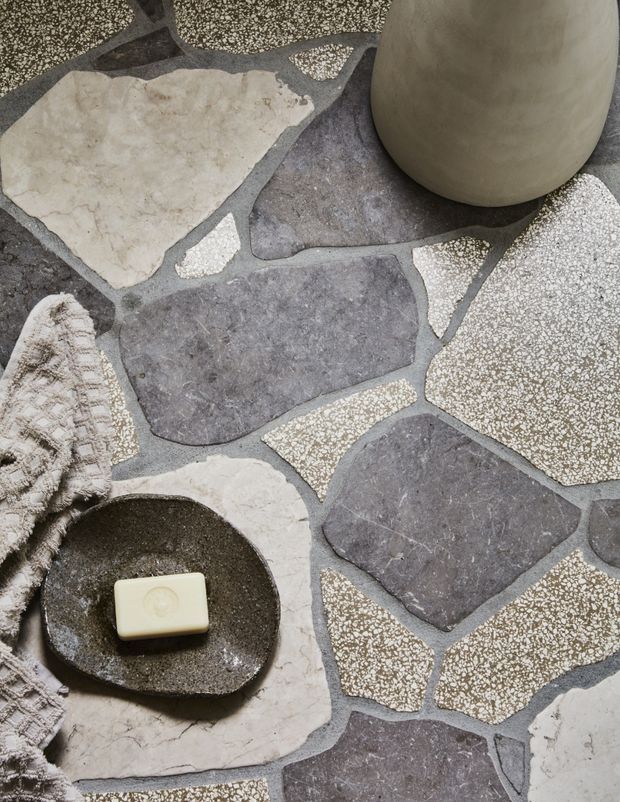 Close-up view of an irregular flagstone floor composed of dark grey, pale stone, and speckled aggregate pieces with contrasting grout lines. A small dark stone dish holding a bar of soap, a neutral ceramic vessel, and a textured woven textile add spa-like styling. The image emphasizes natural, tactile materials and an earthy neutral palette.