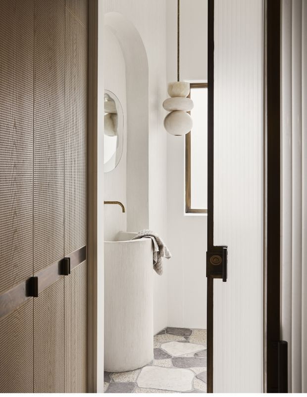 A softly lit, refined bathroom viewed through a partially open dark-framed glass door. The room features a freestanding rounded white basin, a sculptural pendant or wall light with stacked organic forms, a small brass-framed window, white textured walls, and an irregular light stone mosaic floor. A woven textured screen or wall panel occupies the left foreground.