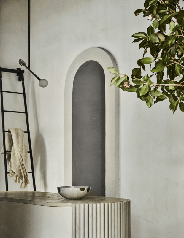 A softly lit, sculptural interior vignette featuring a curved fluted built-in counter or vanity in a pale neutral finish, a tall arched recessed niche with a dark grey interior, and a black leaning ladder with a draped textile. A leafy indoor tree overlaps the upper right of the frame, while a small ceramic bowl sits on the counter. The overall composition is minimalist, tactile, and serene, with strong curved and vertical architectural details.