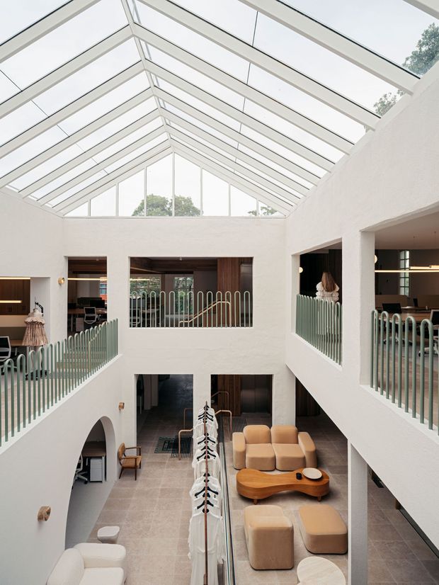 A bright, multi-level interior atrium with a large glazed pitched roof and full-height clerestory glazing. White plaster walls surround an open central void with curved vertical metal balustrades, upper-level openings, and views into adjoining work and lounge areas. The lower level contains a warm-toned lounge arrangement with rounded beige seating, a sculptural timber coffee table, and tiled flooring. The space has a refined contemporary design with strong natural light and indoor greenery visible through the glazing.