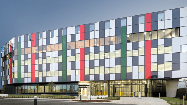 Exterior view of a large contemporary multi-storey commercial building with a gently curved facade composed of a regular grid of glazed panels and opaque cladding panels in white, grey, red, and green. The ground level has extensive glazing, a recessed entrance, and a covered forecourt with vertical metal screening. The building sits along a paved urban street with landscaped planting at its frontage.