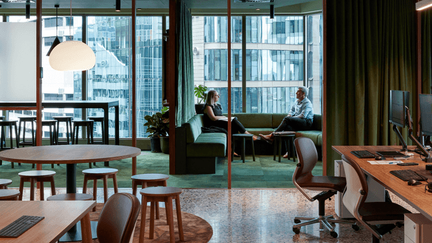 A contemporary commercial office interior with an open-plan collaborative workspace, timber tables and stools, a green upholstered lounge seating area, indoor plants, exposed ceiling services, and workstations with computers. Full-height glazing overlooks dense urban buildings. The space combines warm timber, green textiles, dark curtains, and a polished concrete or terrazzo-like floor with a relaxed hospitality-inspired office layout.