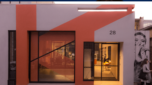 Street-facing facade of a contemporary commercial or retail building, featuring bold geometric red-orange and pale grey wall planes, black-framed glazed openings, a recessed illuminated entrance marked with the number 28, and a large monochrome mural on the adjoining wall. Warm interior lighting is visible through the glazing.