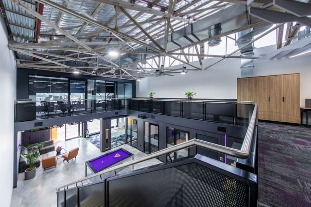 A contemporary multi-level commercial interior viewed from an upper landing, featuring an open mezzanine, exposed steel roof structure and services, extensive glazing, dark metal balustrades, grey flooring, and a bright lower lounge or recreational area with a purple pool table. The space has an industrial office or workplace character with high ceilings and abundant daylight.