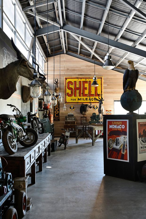 A high-ceilinged industrial-style exhibition space or showroom displaying vintage motorcycles, automotive memorabilia, mounted taxidermy, metal tools, and historic advertising posters. The space features exposed steel roof trusses, corrugated metal roofing, suspended industrial pendant lights, large glazed windows, a polished concrete floor, and a timber-lined feature wall.