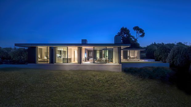 A single-storey modern residence photographed at dusk, with a long, low flat roof, extensive floor-to-ceiling glazing, dark slender window frames, and warm interior lighting. The house opens onto a broad lawn and is partially screened by dense landscaping, creating a strong indoor–outdoor connection.