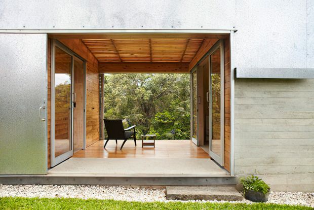 Exterior view of a modern single-storey pavilion-like building with a wide open glazed entrance, warm timber-lined ceiling and walls, and a small furnished sitting area opening toward dense greenery. The facade combines pale textured cladding with concrete elements, timber framing, and expansive folding or hinged glass doors.