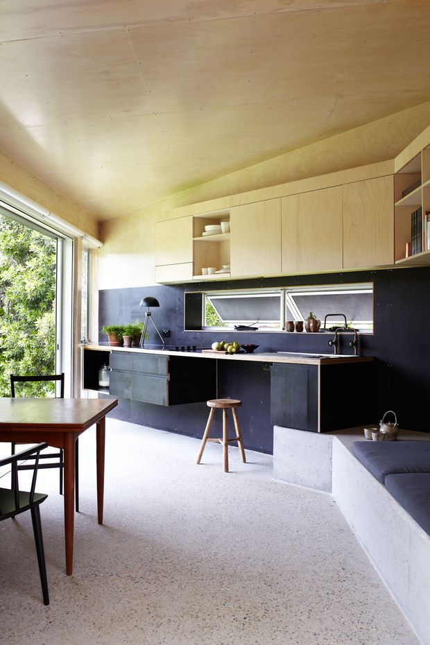 Bright open-plan kitchen and dining area with extensive pale timber wall and ceiling lining, matte black lower cabinetry and backsplash, narrow horizontal awning windows, and a polished concrete or terrazzo-like floor. A timber dining table and chairs occupy the foreground, while a built-in concrete bench with dark cushion seating is visible at right. The space has a simple, functional contemporary character with strong natural light and garden views through large glazing.