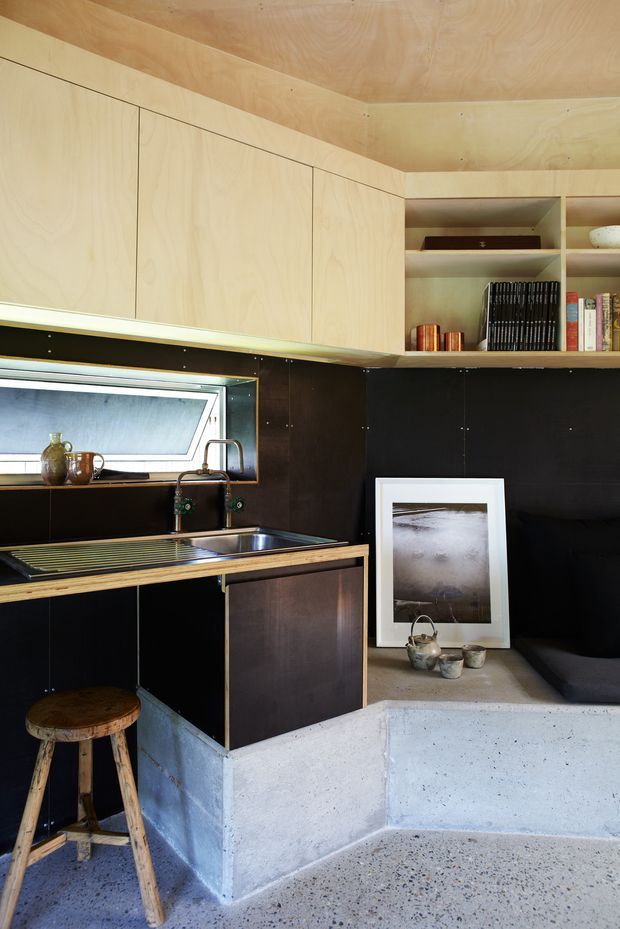 Compact, highly integrated kitchen corner with pale plywood overhead cabinetry and ceiling, dark wall panels, open shelving, a stainless-steel sink and draining board beneath a horizontal window, and a small timber island or counter. A raised concrete-plinth seating nook and terrazzo-like floor add a raw, utilitarian character. The space is styled with books, ceramics, framed artwork, and a timber stool.