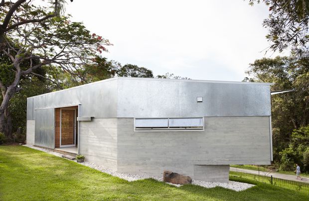 A compact single-storey modern concrete dwelling set within a leafy landscaped site. The building has a flat roof, smooth pale concrete walls, a silver-grey metal-clad side volume, a timber entry door, narrow horizontal windows, and a partially elevated concrete form over a gravel strip.