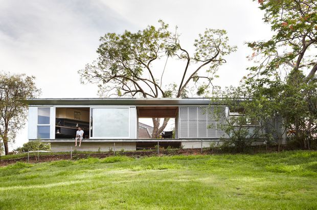 A low, single-storey modern house viewed from the garden, with a long horizontal composition, flat roof, extensive sliding glazing, pale cladding, and a screened central opening. Mature trees surround the building and a gently sloping lawn occupies the foreground.