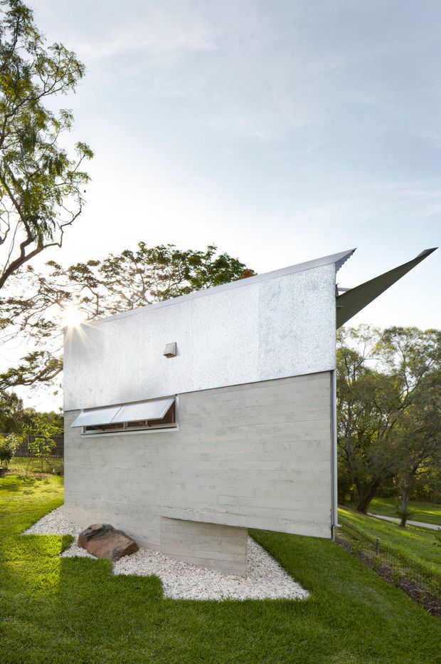 Exterior view of a compact, sculptural modern building set within a landscaped grassy site. The facade combines pale textured rendered upper walls with horizontal concrete or cement-board-like lower cladding, a narrow projecting window with an external awning, and sharply angled roof or canopy forms extending beyond the main volume. White gravel borders the building, with mature trees and a sloping garden surrounding it.