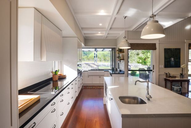 Bright, elongated contemporary kitchen with extensive white built-in cabinetry, dark benchtop accents, a large pale island with integrated sink, polished timber flooring, and a coffered ceiling. Pendant lights hang above the island, while glazed doors at the rear connect visually to an outdoor area. A furnished living space is visible beyond the kitchen.