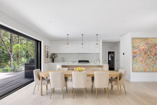 Bright open-plan dining and kitchen interior with a large light timber dining table surrounded by upholstered chairs, a white vertically panelled kitchen with island, pale timber flooring, pendant lights, extensive black-framed glazing, and a colourful wall artwork. Lush trees are visible outside through the glazing.