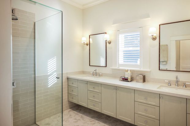 Bright, spacious traditional bathroom featuring a long double vanity with pale grey-green shaker cabinetry, light stone benchtop, twin undermount basins, framed mirrors, polished chrome fixtures, and a large frameless glass walk-in shower. The shower has light grey subway wall tiles, while the floor features pale patterned tiles. White walls, plantation-style window shutters, and classic wall sconces create a clean, refined atmosphere.