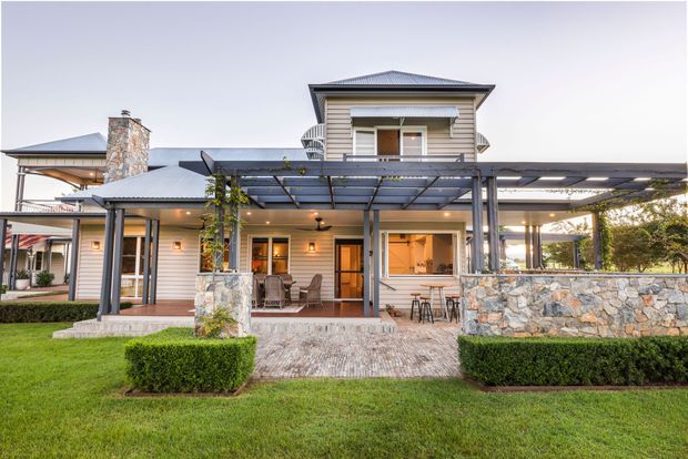 Exterior view of a substantial multi-storey home with a light weatherboard facade, dark hipped metal roof, broad covered outdoor entertaining area, timber-framed glazing and doors, and prominent natural stone chimney and retaining wall elements. A landscaped lawn, clipped hedges, paved terrace, and pergola-style roof structure frame the house.