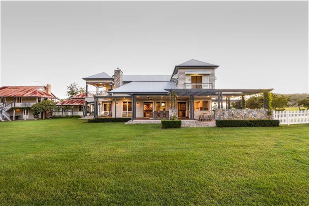 Exterior view of a large multi-level home set behind an expansive lawn. The house features a light rendered facade, dark metal roofing, multiple gabled roof forms, covered verandas and patios, extensive glazing, timber-framed openings, stone-clad sections, and a long pergola-like covered outdoor area. A neighbouring or connected building with weathered red metal roofing is visible to the left, along with a white picket fence and landscaped hedges.