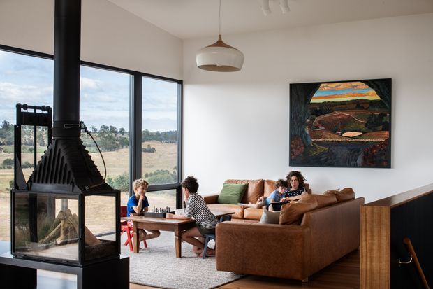 A bright, modern living room with expansive floor-to-ceiling glazing overlooking a rural landscape. A freestanding black wood-burning fireplace forms a strong focal point beside a large brown sectional sofa, with children seated at a timber coffee table. White walls, timber flooring, a sculptural pendant light, and a colourful framed artwork complete the warm contemporary interior.