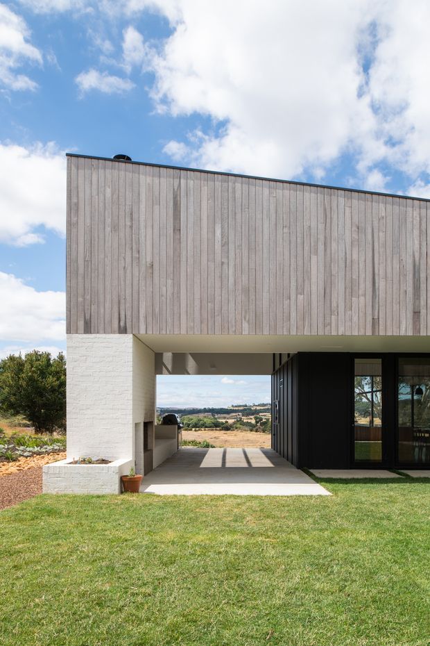Exterior view of a contemporary single-storey residence with a long, flat-roofed form. The upper facade is clad in vertically arranged weathered timber boards, while the lower level combines white-painted brick, a deep covered outdoor passage, and dark-framed glazed doors. The house opens toward a landscaped lawn and a broad rural valley beyond.