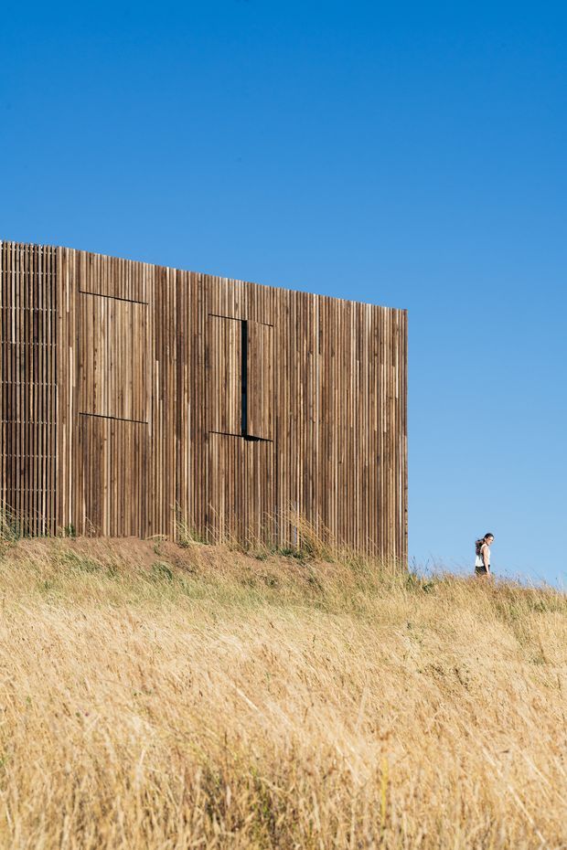 Exterior architectural photograph of a minimalist rectangular building partially embedded in a dry grassy slope. The facade is clad in closely spaced vertical timber battens, with narrow recessed openings and a simple sloping roofline or upper edge. A person provides scale, while the clear blue sky and surrounding grass emphasize the isolated landscape setting.