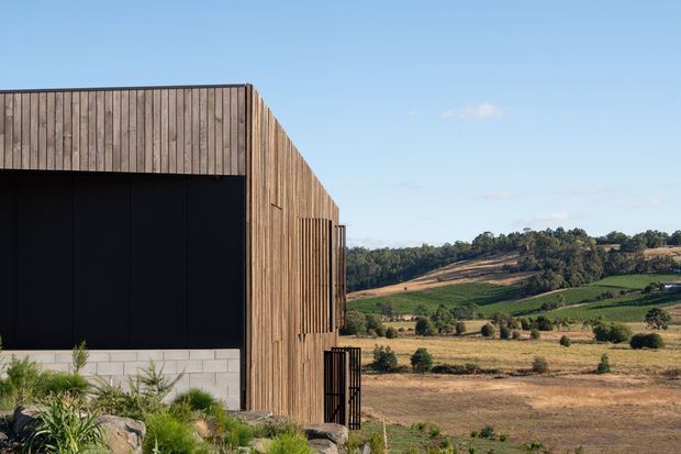 Exterior view of a contemporary rural building with a strongly rectilinear form. The facade combines vertical timber cladding with a large matte-black recessed opening and a low pale masonry base. Native-style landscaping sits in the foreground, while rolling cultivated countryside is visible beyond under a clear blue sky.