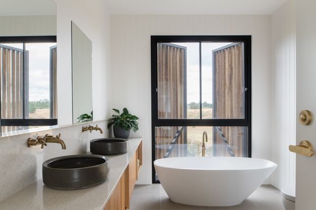 Bright contemporary bathroom featuring a freestanding white oval bathtub, two dark vessel basins on a pale stone vanity, brushed brass wall-mounted tapware, timber cabinetry, vertical light wall panelling, and black-framed glazing with exterior timber screening. A large mirror reflects the vanity and window.