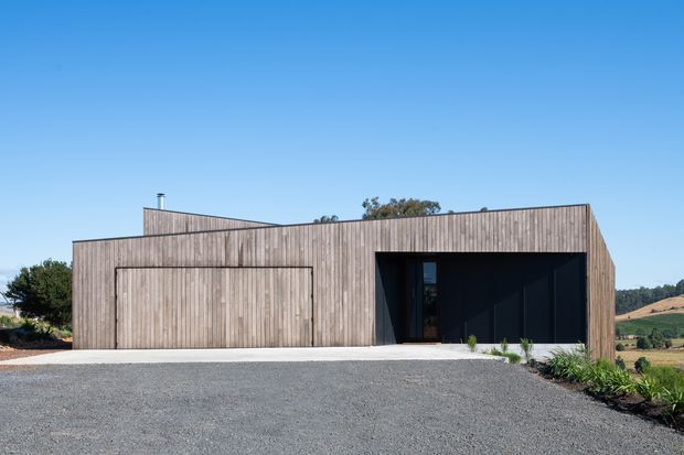 A minimalist single-storey building exterior in a rural landscape, clad in vertical natural timber boards. The angular facade includes a large flush garage or service door on the left and a deep, dark recessed entry or covered opening on the right. A broad gravel driveway occupies the foreground, with sparse planting along the building edge and clear blue sky above.