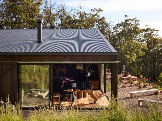 A compact single-storey timber-clad cabin set within a wooded landscape. The front facade features broad glazing and sliding doors opening onto a timber deck furnished with outdoor lounge chairs and a small table. A dark corrugated pitched roof with a prominent flue defines the simple rural form, while a landscaped gravel garden and surrounding native woodland are visible around it.