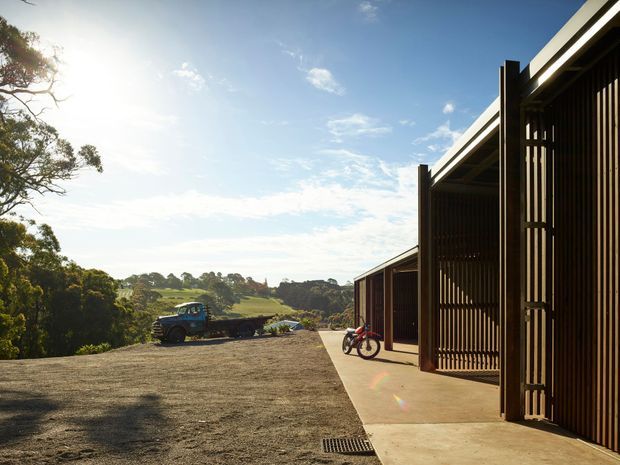 Exterior view of a contemporary dark-clad building with tall vertical slatted screening and a projecting roofline. A broad gravel forecourt and concrete walkway lead along the facade, with bicycles and a utility vehicle visible. The building sits in a leafy, gently rolling rural landscape under bright sunlight.