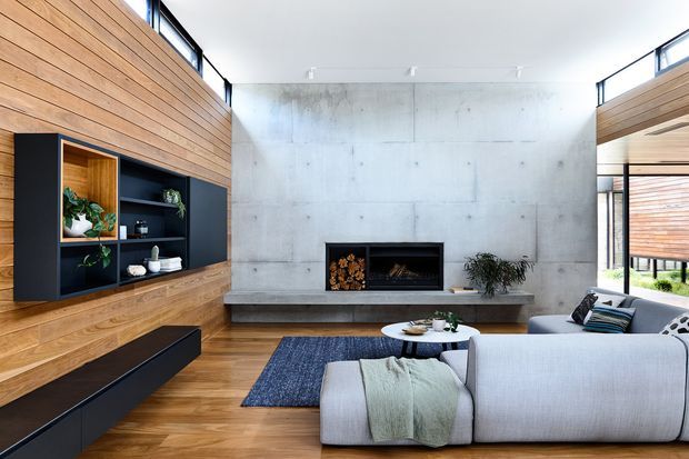 Bright contemporary living room with full-height concrete fireplace wall, horizontal timber wall panelling, integrated dark built-in shelving and low cabinetry, and a large pale sectional sofa arranged around a round coffee table. Expansive glazing and clerestory windows bring in natural light, with a view toward an outdoor area on the right.