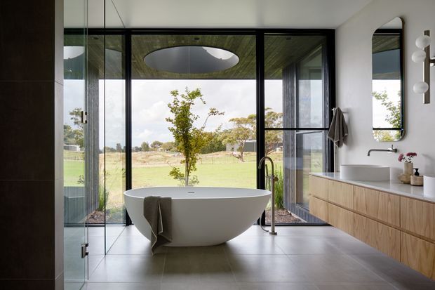 A contemporary, light-filled bathroom with a freestanding white oval bathtub positioned in front of full-height black-framed glazing. The room features large-format grey floor tiles, a timber floating vanity with white countertop and integrated basins, a rounded wall mirror, and a wall-mounted sculptural light. A landscaped rural outlook is visible through the glazing, while a skylight or roof opening adds overhead illumination.