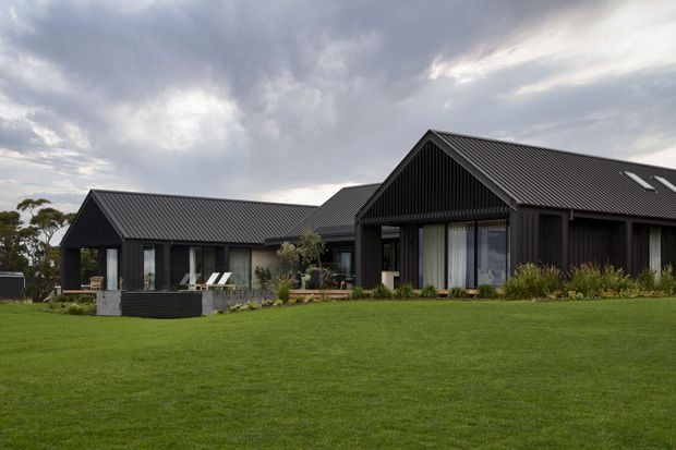 A single-storey contemporary residential home set within a broad lawn. The building has multiple pitched gable roof volumes, dark ribbed metal roofing, dark vertical cladding, extensive floor-to-ceiling glazing, and a covered outdoor-facing veranda or entry zone. Low landscaping and a landscaped retaining planter sit near the facade beneath an overcast sky.