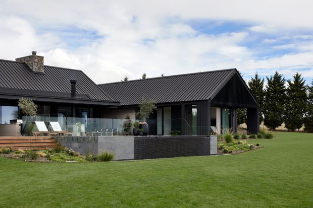 Exterior view of a contemporary single-storey home with dark corrugated metal roofing and vertical dark cladding, a prominent gabled wing, large glazed openings, and a timber deck with glass balustrade. The house is set within a broad manicured lawn and landscaped garden, with a stone chimney and mature screening trees.