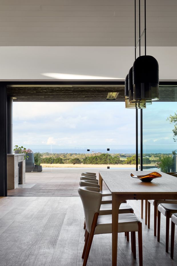 A refined dining area opening onto a broad terrace with expansive ocean and coastal views. The room features a long light-wood dining table, upholstered dining chairs, sculptural black pendant lights, dark-framed floor-to-ceiling glazing, and a pale linear-panel ceiling. A fireplace or built-in feature is visible to the left, while the terrace and landscaped coastal setting extend beyond.