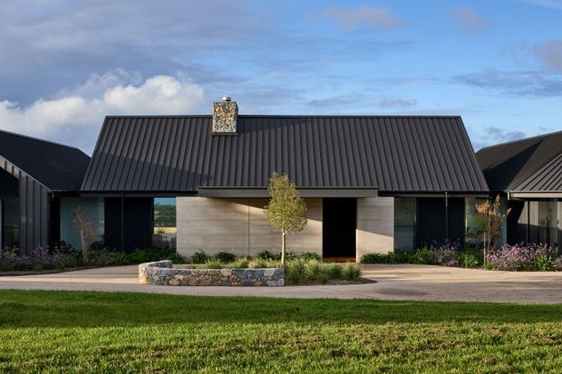 Single-storey contemporary residential building viewed from the front, with a steeply pitched dark metal gable roof, pale rendered or concrete-toned facade, dark vertical wall sections, broad glazing, and a central entry recessed beneath a projecting roof edge. A stone-edged garden bed and lawn occupy the foreground, while attached dark-roofed wings extend on both sides.
