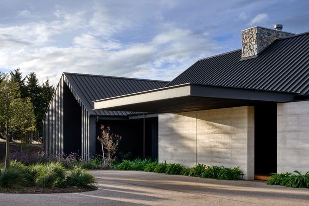 Exterior view of a contemporary single-storey residence with steeply pitched black ribbed metal roofs, dark vertical cladding, pale textured concrete walls, and a projecting flat-roofed entry volume. A natural stone chimney rises above the roofline, with landscaped planting and a paved approach in the foreground.