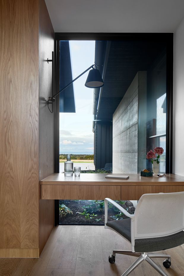 Compact built-in home office nook with a wall-mounted desk spanning a large framed opening. The space features warm wood cabinetry and desk surfaces, a black articulated wall lamp, pale timber flooring, a white ergonomic office chair, and a strong view through the opening toward an outdoor courtyard and distant landscape. A textured concrete wall and dark roof structure are visible beyond.