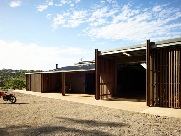 A low-profile contemporary building viewed from a broad gravel forecourt. The structure features dark timber vertical batten screens, large open garage-like bays, a flat or very low-pitched roof, and simple concrete paving. Rolling bush-covered terrain is visible beyond the building under a bright partly cloudy sky; a motorcycle is parked at the edge of the frame.