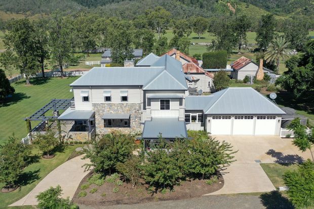 Aerial view of a substantial rural residential property with a light grey metal roof, pale stone-clad walls, attached double garage, paved driveway, landscaped garden, and several adjoining or nearby outbuildings. Rolling green hills and mature trees form the background.