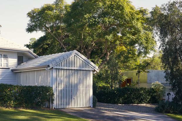 Exterior view of a residential property featuring a white weatherboard house and a prominent detached garage or carport structure with vertical slatted cladding and a pitched gable roof. Mature trees, hedges, lawn, and a gravel driveway surround the buildings. The scene has a calm, established suburban character with strong daylight and soft natural greenery.